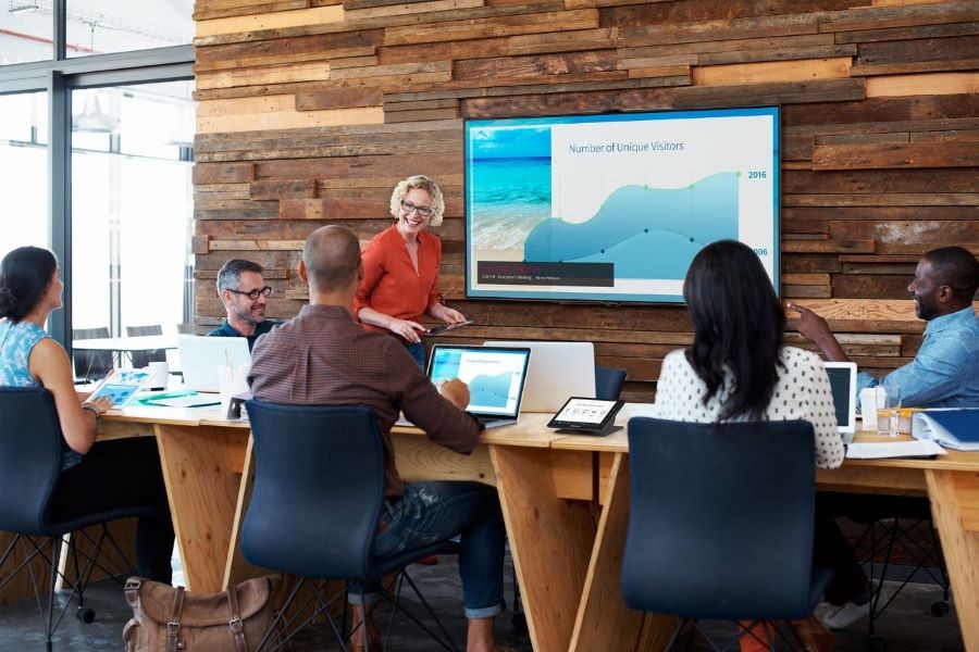 A huddle space with a video display and people sitting around a table using their laptops.