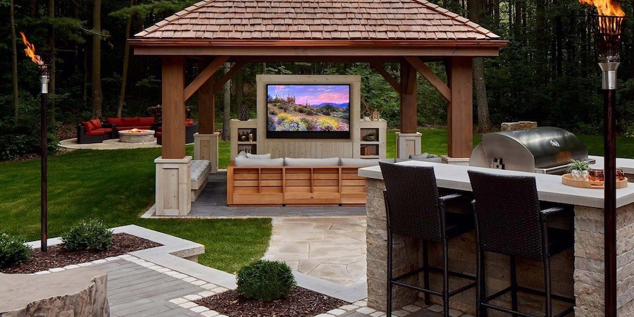 An outdoor kitchen with a TV, grill, and seating, surrounded by greenery