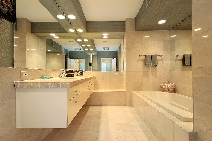 Modern bathroom with beige marble tile, built-in bathtub, floating vanity with stone countertop, large mirrors, and recessed lighting.