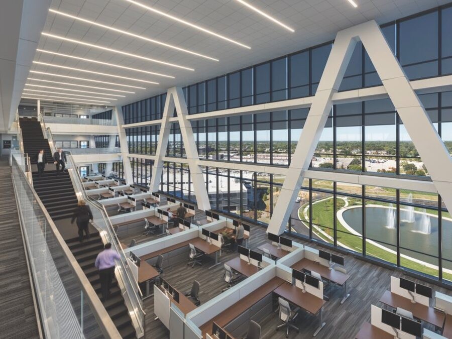 Modern open-concept office space with large floor-to-ceiling windows, rows of desks, and a grand central staircase, offering views of a landscaped area with a pond and fountains.